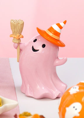 Fantôme Halloween rose pastel tenant un balai et coiffé d'un chapeau orange rayé, sur une table d'Halloween